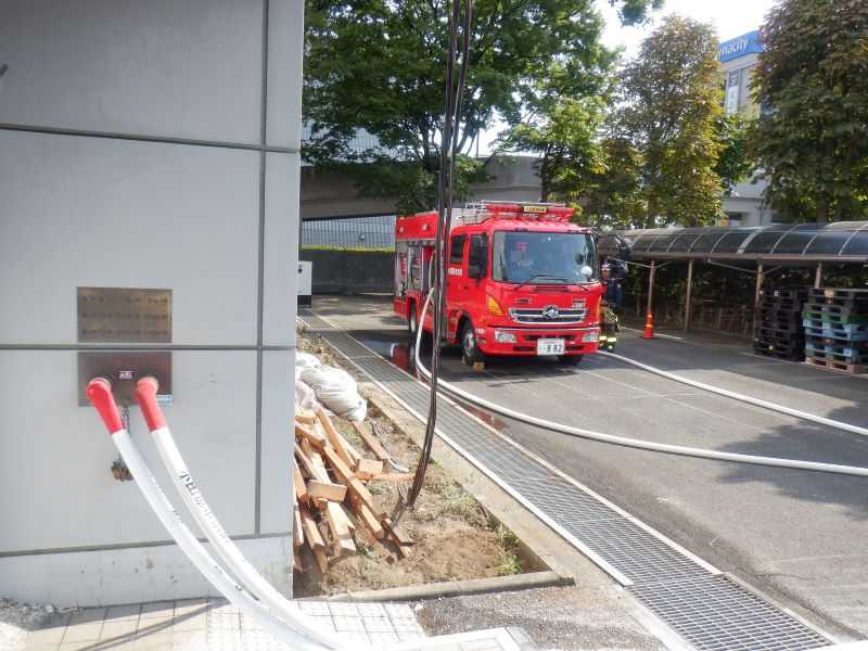 敷地内の防火水槽から消防車で吸水し、建物に設置されている連結送水管へ送水することにより、上層階までホースを延長することなく放水している写真