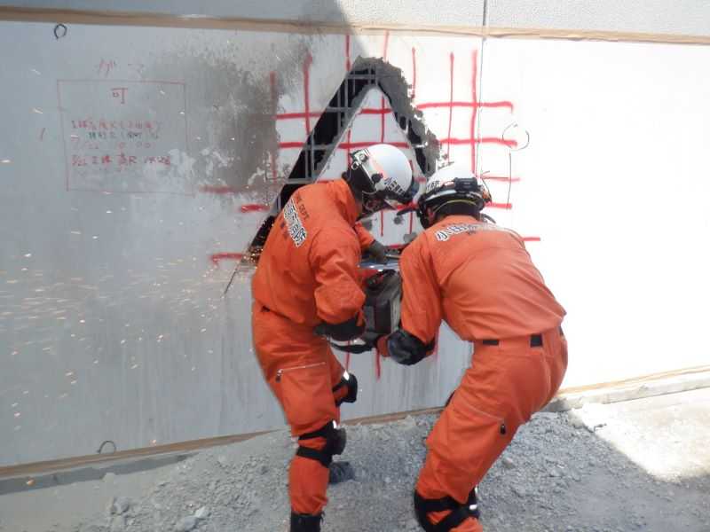 震災等により座屈した建物を想定し、閉じ込められた要救助者を救出するため、削岩機やエンジンカッター等を使用して鉄筋コンクリートの壁を破壊している写真。